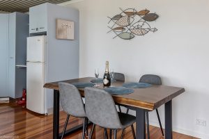 A modern dining area with a wooden table set for two, featuring a bottle of champagne and glasses. Four gray chairs surround the table, and a metal fish wall art decorates the white wall behind. A refrigerator is visible in the background.