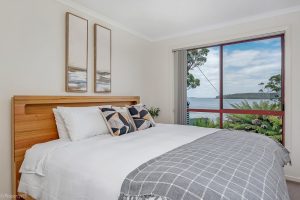 Modern bedroom with a wooden bed, white bedding, patterned pillows, and a gray checked throw, featuring two abstract paintings and a large window with a scenic view of water, trees, and blue sky.