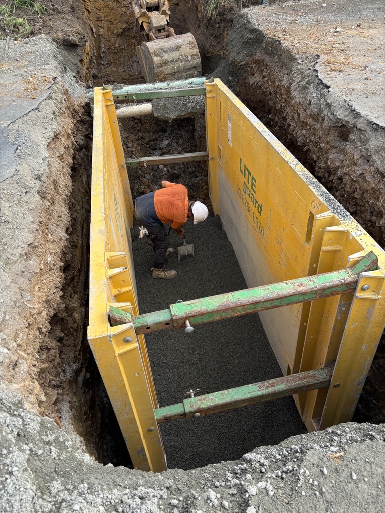 A construction worker in an orange shirt and hard hat uses a shovel to level gravel inside a yellow trench box within an excavation pit.