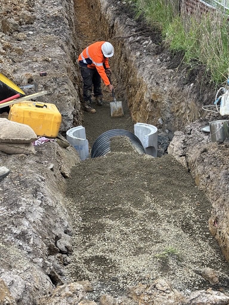A construction worker in a high-visibility jacket and hard hat shovels gravel into a trench containing a large drainage pipe.