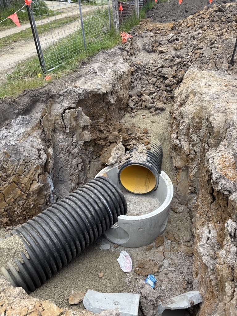 Black corrugated drainage pipes connected to a concrete junction box in an open construction trench.