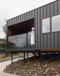 A person in a white robe and towel stands on the balcony of a modern, dark metal house elevated on stilts, looking out over a rocky, sloped landscape.