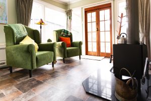 A cozy living room with two green armchairs, colorful cushions, a floor lamp, and large windows with curtains. Sunlight streams in through glass doors, and a wood-burning stove sits in the foreground.