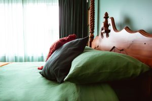 A neatly made bed with green sheets and pillows, a dark gray pillow, and a reddish-orange blanket, beside a carved wooden headboard. Soft light filters through sheer curtains and dark drapes in the background.