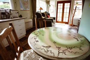 Round wooden table with a painted bird and branch design in a cozy, sunlit living area. Two chairs are nearby, and the room features armchairs, a map on the wall, and glass doors leading outside.