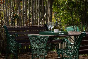 A green metal garden table with ornate chairs holds a tray with a wine bottle and two glasses. A wooden bench and greenery, including trees and a wooden fence, are in the background. Sunlight casts shadows on the scene.