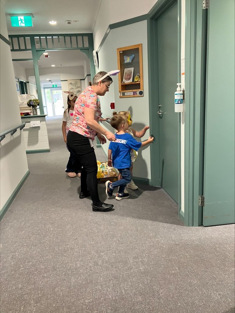 An adult wearing bunny ears and three children walk down a hallway. One child wears a blue shirt and bunny ears and is knocking on a door, while the others carry Easter baskets. The scene suggests an Easter egg hunt indoors.