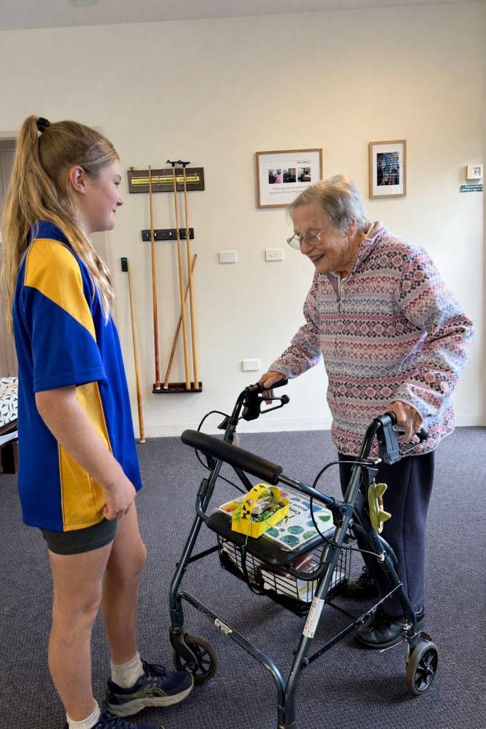 A young girl in a blue and yellow shirt talks to an elderly woman using a walker. The woman is smiling, and there are framed photos and walking sticks on the wall behind them.
