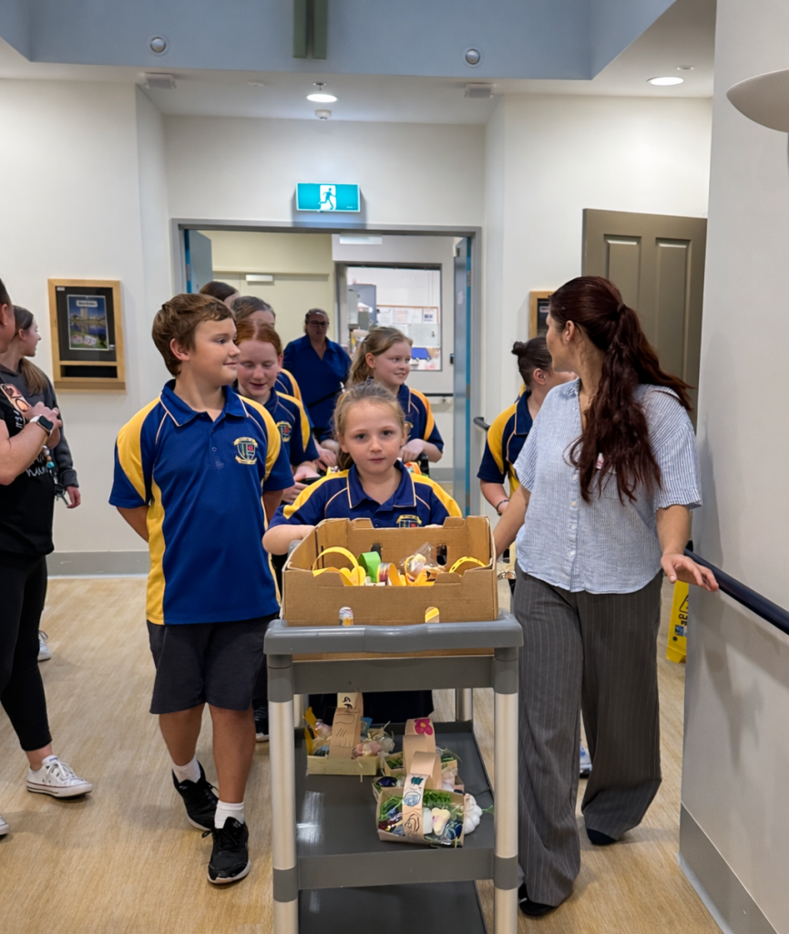 A group of school children in blue and yellow uniforms walk down a hallway with a teacher, pushing a cart filled with snacks and drinks. The setting appears to be indoors at a school or community center.