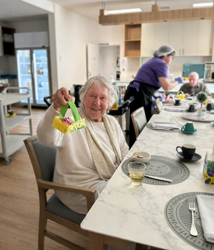 An elderly woman smiles and holds up a colorful handmade basket while sitting at a dining table in a bright, modern room. Other people and a staff member are present in the background.