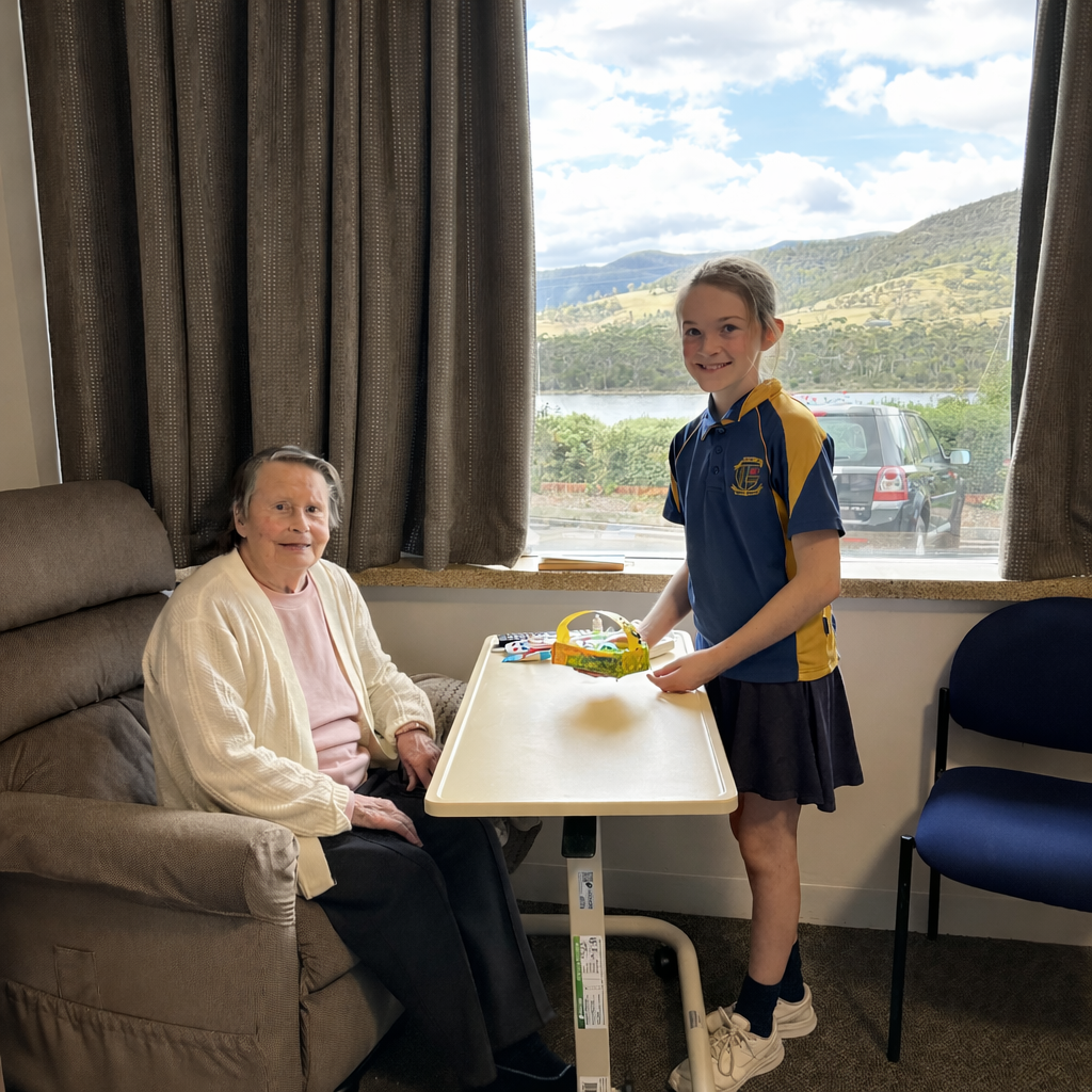 An elderly woman sits in a chair by a table, smiling at a young girl in a school uniform holding a basket. They are indoors with a window view of green hills and a partly cloudy sky in the background.