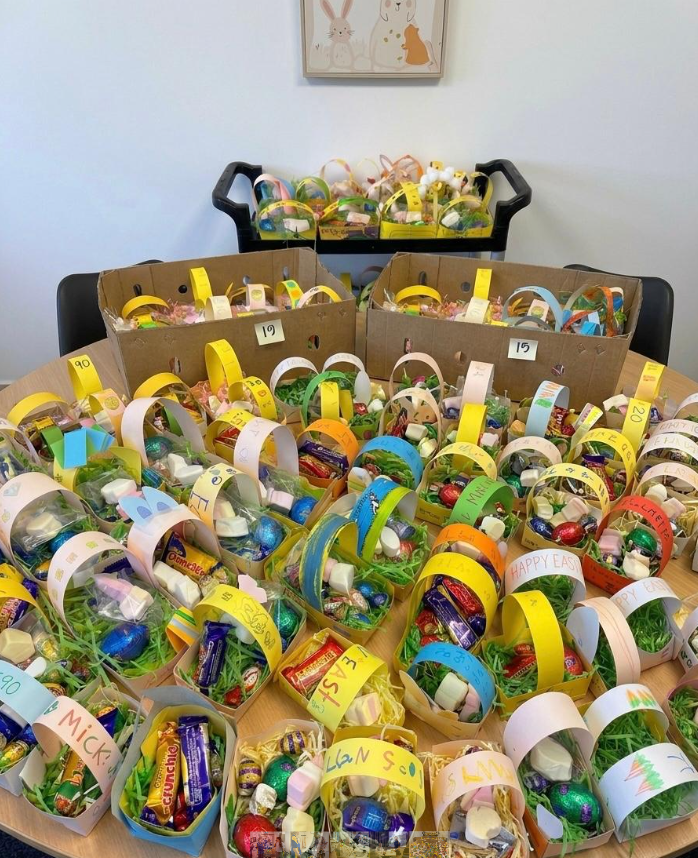 Colorful handmade Easter baskets filled with chocolate eggs, candy, and treats are arranged on a table and a cart. Each basket has a yellow paper handle labeled with a different name. A framed animal print hangs on the wall.
