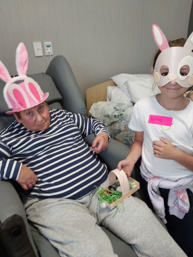 An elderly man with a pink bunny hat sits in a chair next to a young girl wearing a bunny mask and holding a basket with Easter grass and a bunny cutout. Both appear to be participating in an Easter activity.