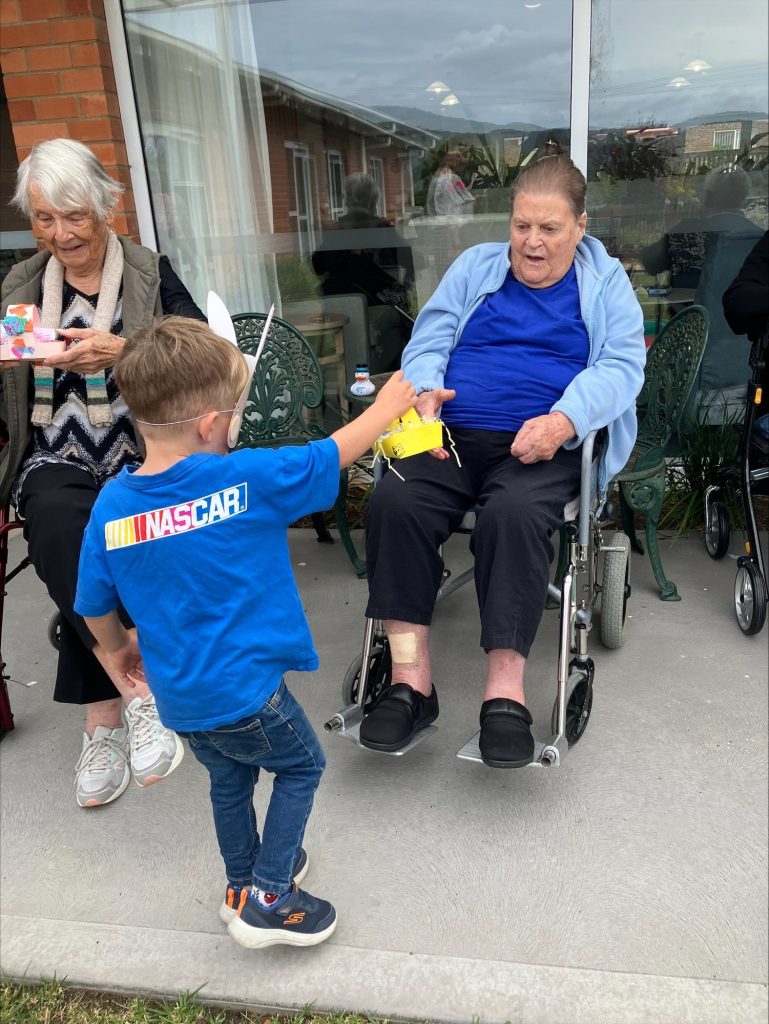 A young boy hands a yellow object to an elderly woman in a wheelchair outside, while another elderly woman sits nearby holding a colorful box. They are on a patio in front of a building with large windows.