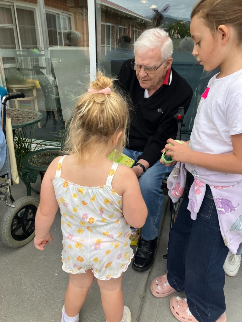 An elderly man sitting outside interacts with two young girls. One girl faces away, wearing a white floral outfit, while the other stands nearby holding a green object. A walker and patio furniture are in the background.
