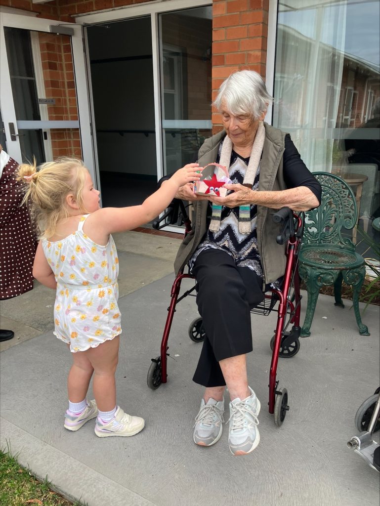 A young girl hands a craft heart to an elderly woman sitting on a walker chair outside a building. Both are smiling and interacting warmly.