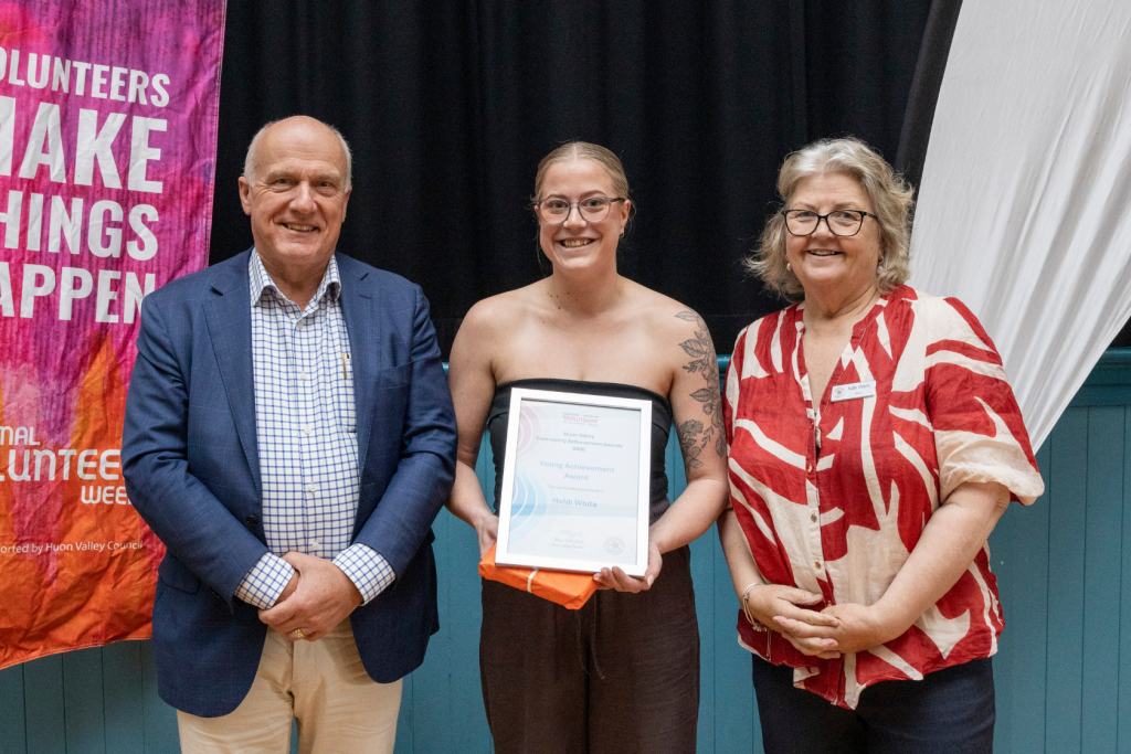 Three people stand for a photo. A man in a blue suit and plaid shirt on the left, a young woman holding an award in the middle, and a woman in a red and white patterned shirt on the right.
