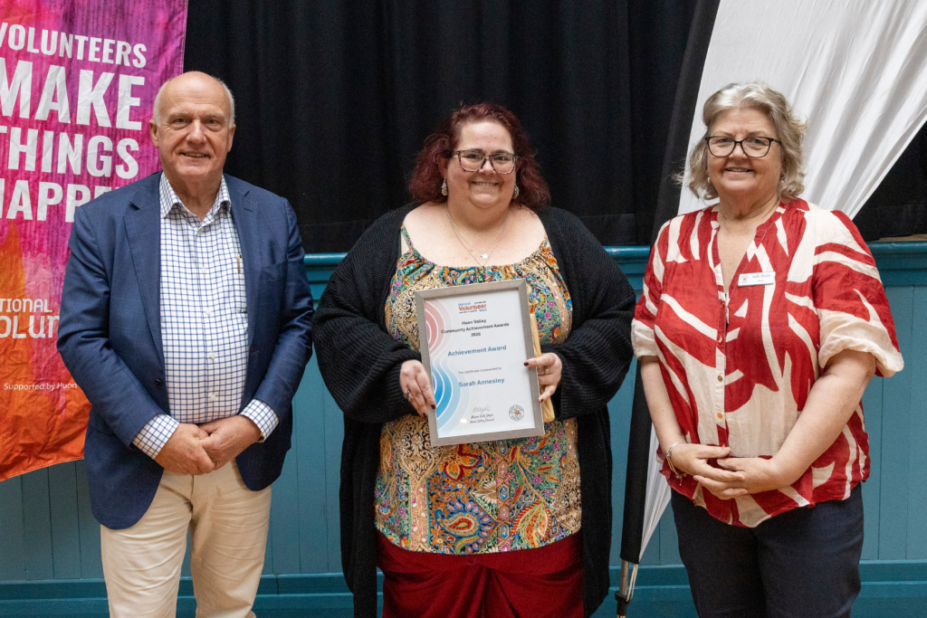 Three people stand for an award ceremony. The woman in the middle holds an award for Sarah Annesley.