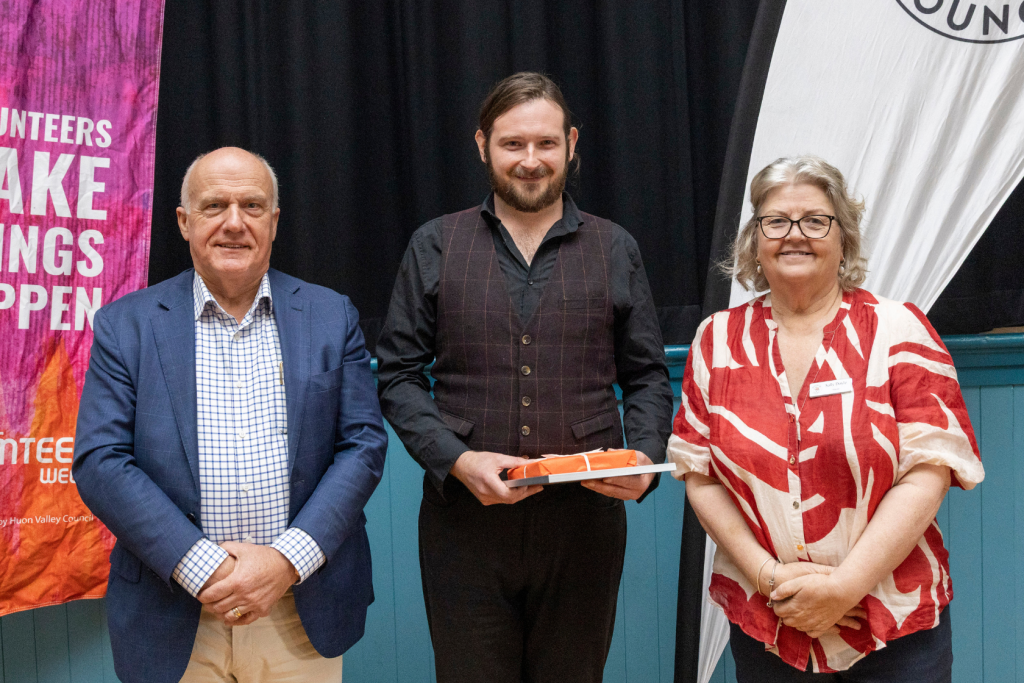 Three people pose for a photo. On the left, a man in a blue suit and checkered shirt smiles. In the middle, a man with a beard in a vest and black shirt holds an orange object. On the right, a woman in a red and white patterned shirt smiles.