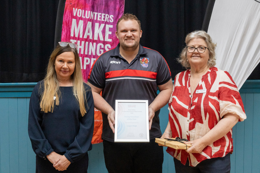 Two women and one man stand in front of a banner. The man in the center holds a framed award.