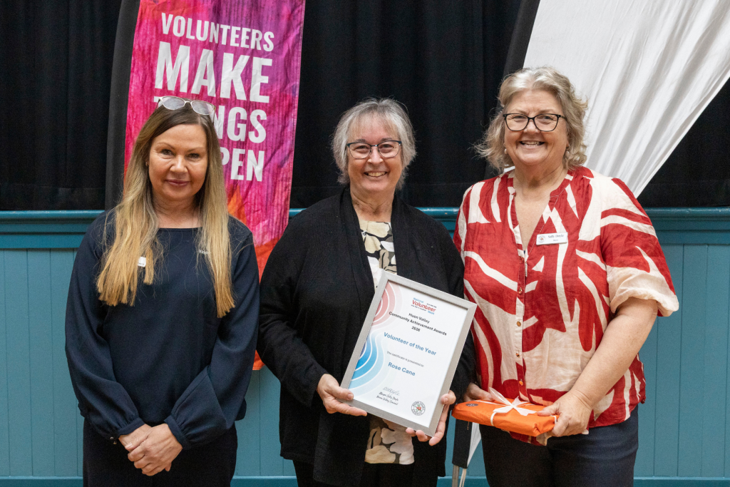 Three women stand for a photo. The woman in the center holds a certificate titled Volunteer of the Year.