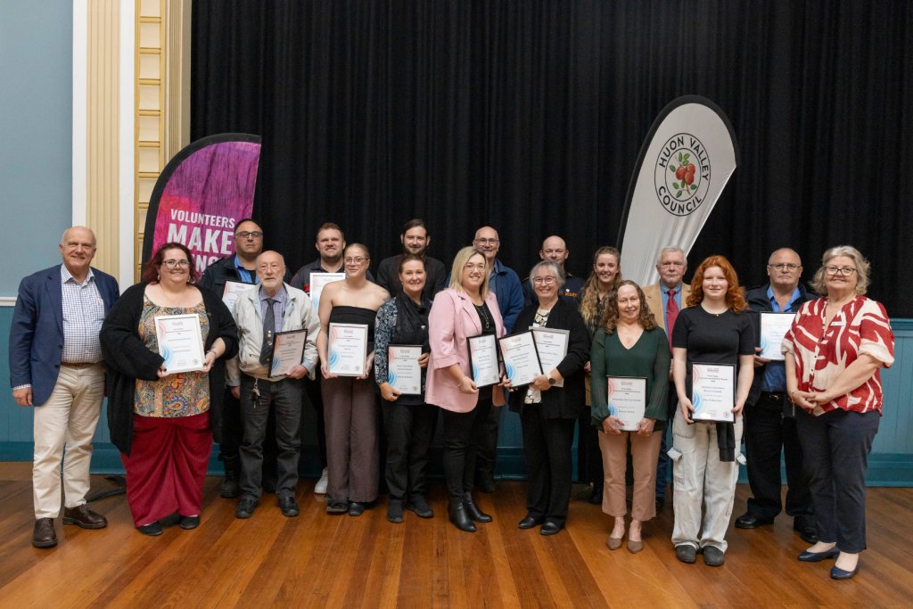 A group of people, mostly women, stand in front of a black curtain holding certificates. A banner behind them reads VOLUNTEERS MAKE.