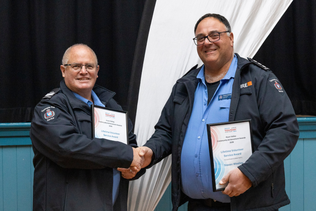 Two men in Tasmania Fire Service jackets shake hands, each holding a framed certificate for a Lifetime Volunteer Service Award.