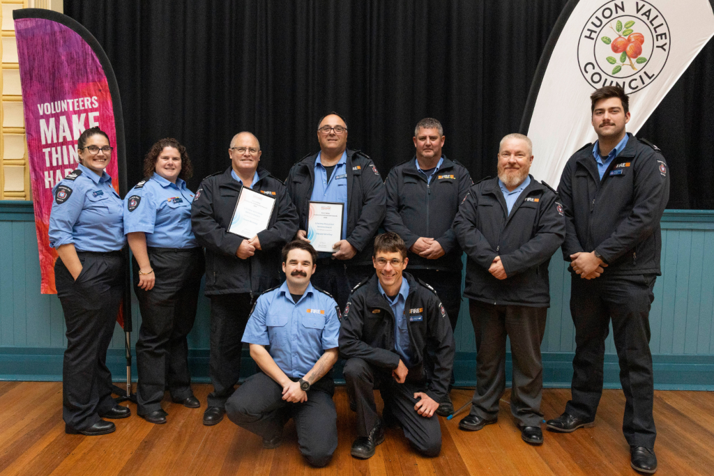 Group photo of 9 firefighters in uniform, some holding certificates, with a Huon Valley Council banner in the background.