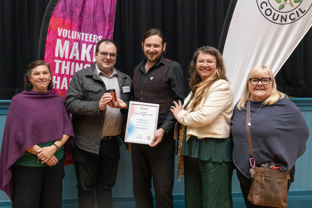 Five people stand together holding an award. The person in the center smiles while holding a certificate. The others smile and one person holds a jar.