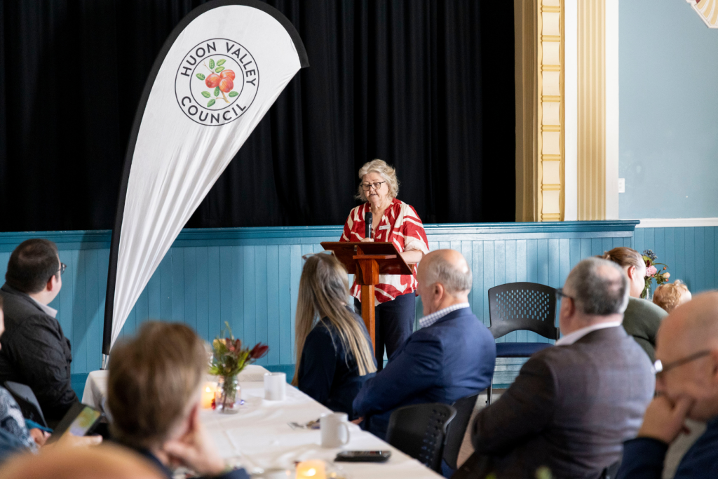 A woman in a red and white patterned shirt speaks at a podium in front of a banner for the Huon Valley Council. An audience is seated facing her.
