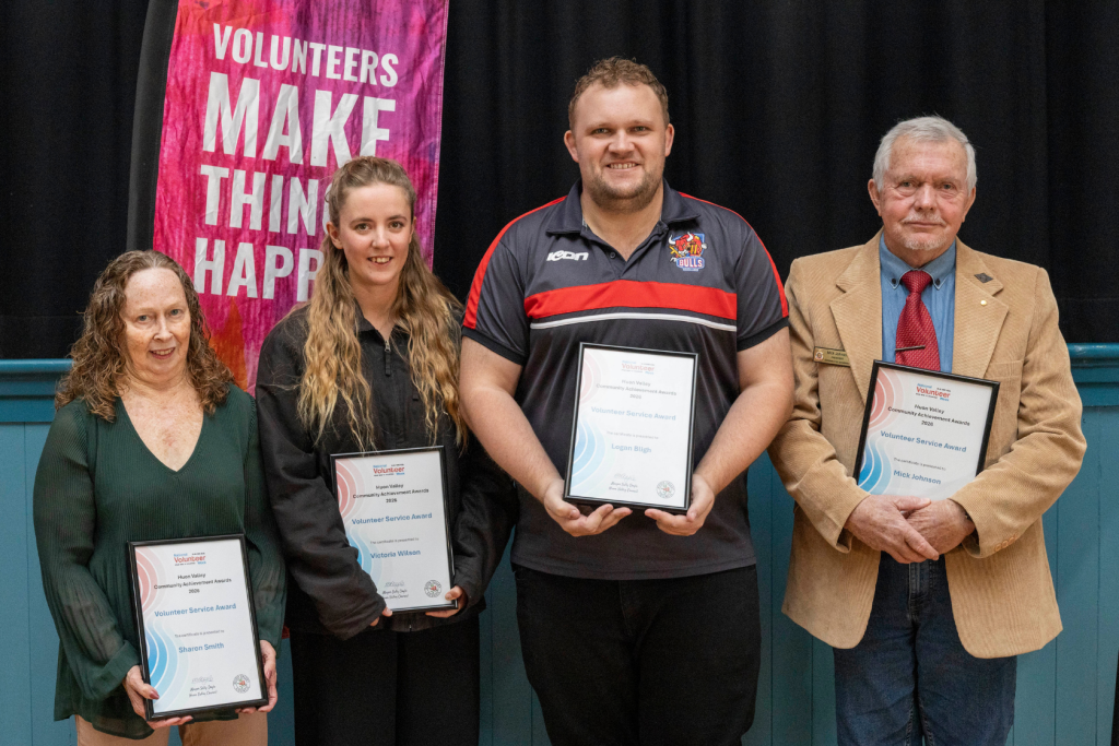 Four volunteers, Sharon Smith, Victoria Wilson, Logan Bligh, and Mick Johnson, are presented with Volunteer Service Awards in front of a Volunteers Make Things Happen banner.