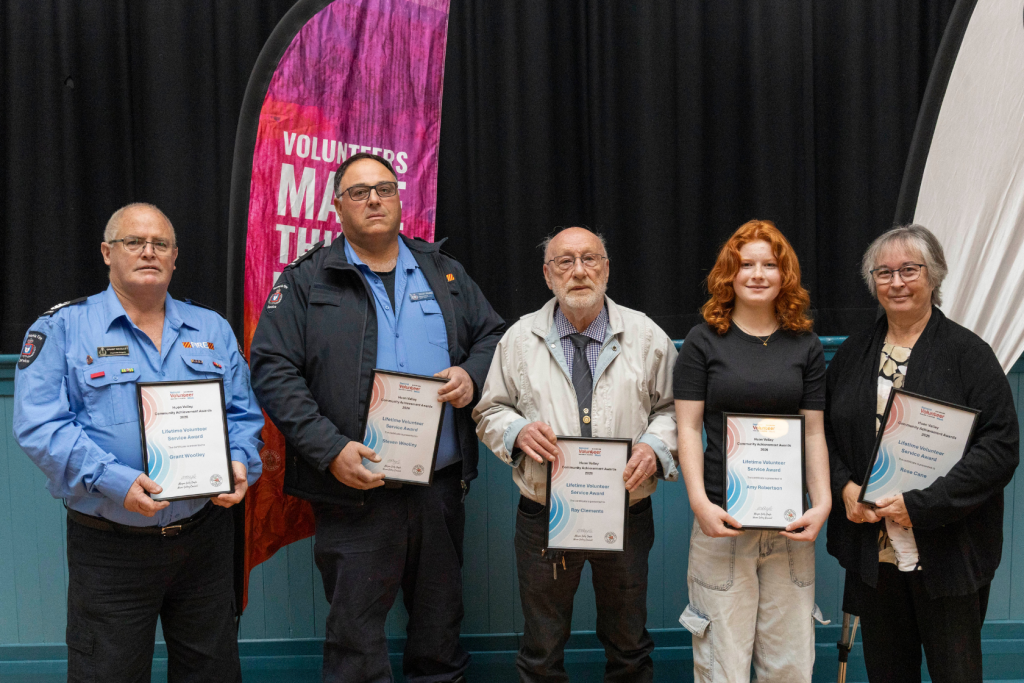 Five people, four with awards, stand in front of a black curtain and a pink banner. The people are Grant Woodley, Steven Woolley, Ray Clements, Amy Robertson, and Rose Carle.