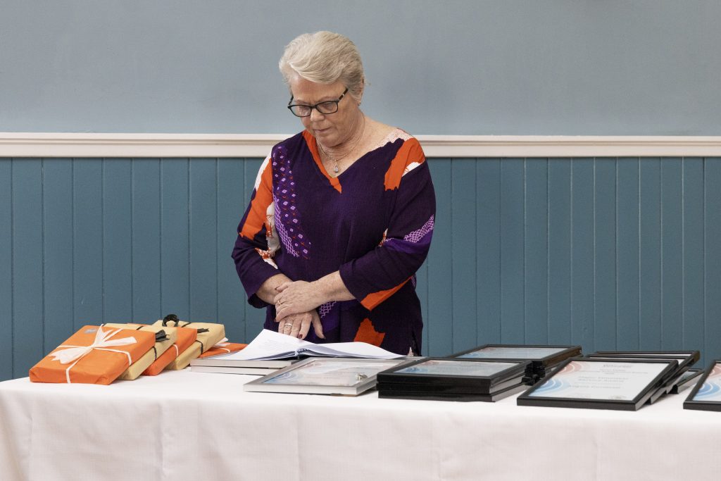 An older woman with glasses, wearing a purple and orange patterned shirt, looks down at a book on a table. Gifts are on her left, and framed certificates are on her right.