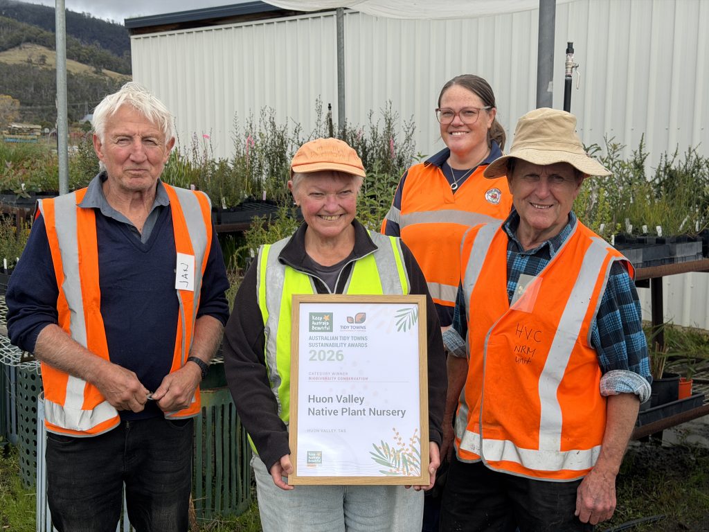 Four smiling people in orange safety vests stand outdoors near potted plants. The central person holds a framed certificate that reads Huon Valley Native Plant Nursery 2026. A shed and hills are visible in the background.