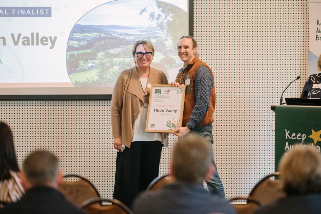 Two people stand smiling and holding a framed award certificate labeled Huon Valley at a presentation event, with a large screen and audience members visible in the background.