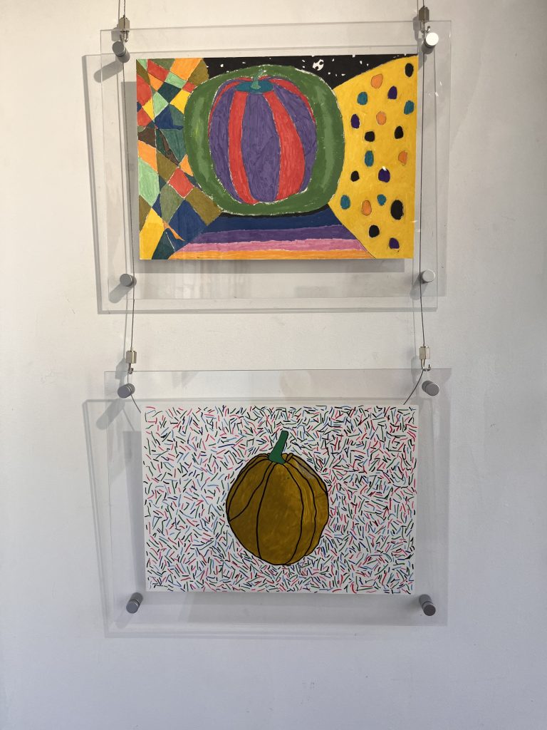 Two paintings of pumpkins are displayed on a wall. The top painting features a colorful, geometric background, while the bottom painting shows a pumpkin with a white background covered in red and green sprinkles.