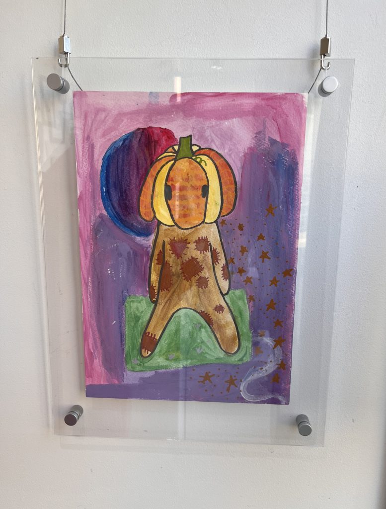 A colorful painting of a figure with a pumpkin for a head, standing on green ground. The background is purple and pink with a blue circle and orange stars scattered on the right side. The artwork is framed in clear acrylic.