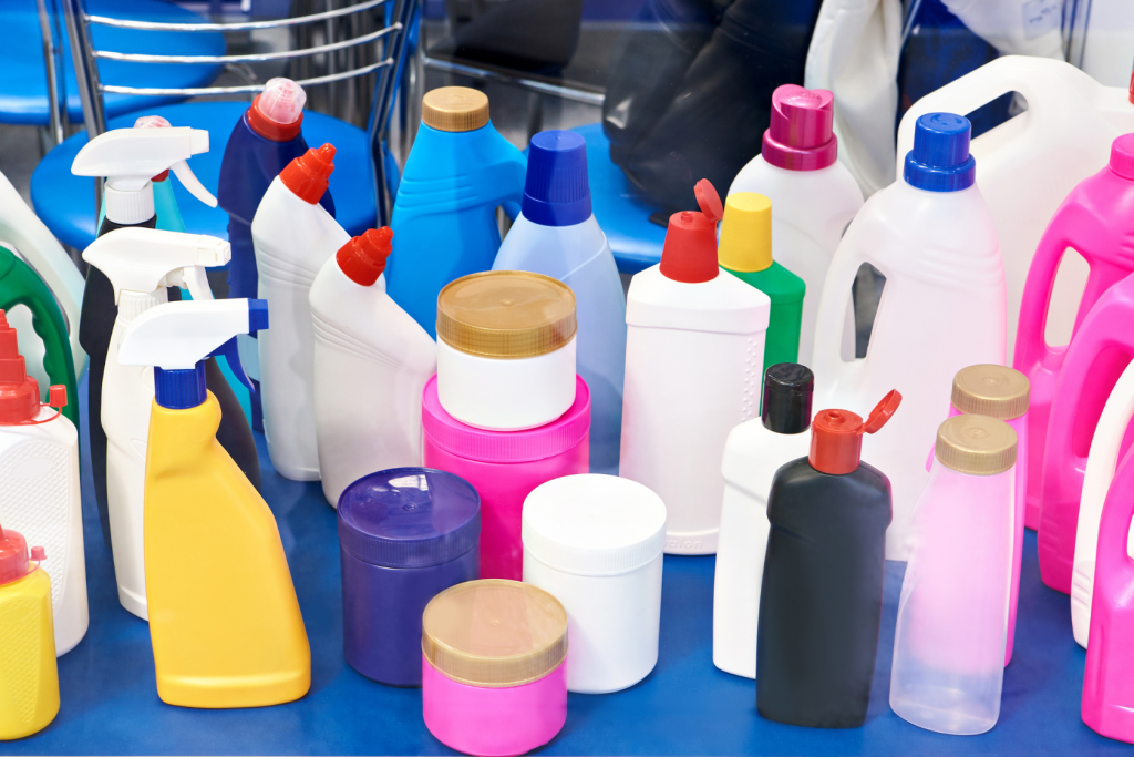 A variety of colorful plastic bottles and containers, including spray bottles, detergent bottles, and jars, are displayed on a blue surface. The containers have different shapes, sizes, and colored caps.