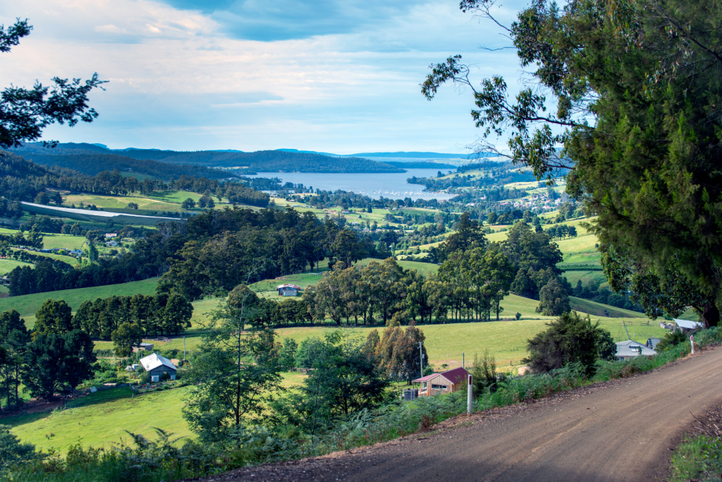 A rural landscape with green fields, scattered houses, and trees, overlooking a lake in the distance. A dirt road runs along the right side, bordered by lush vegetation under a blue sky with light clouds.