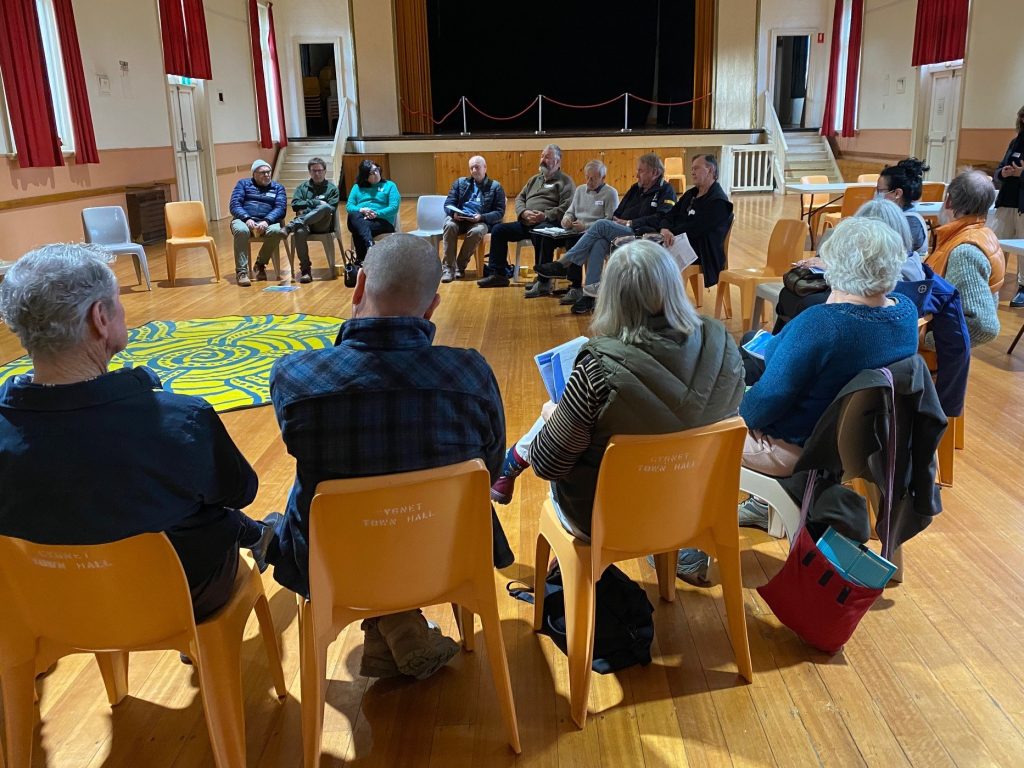A group of people sit in a large circle on yellow chairs in a hall with wooden floors and red curtains, engaged in a discussion or meeting.