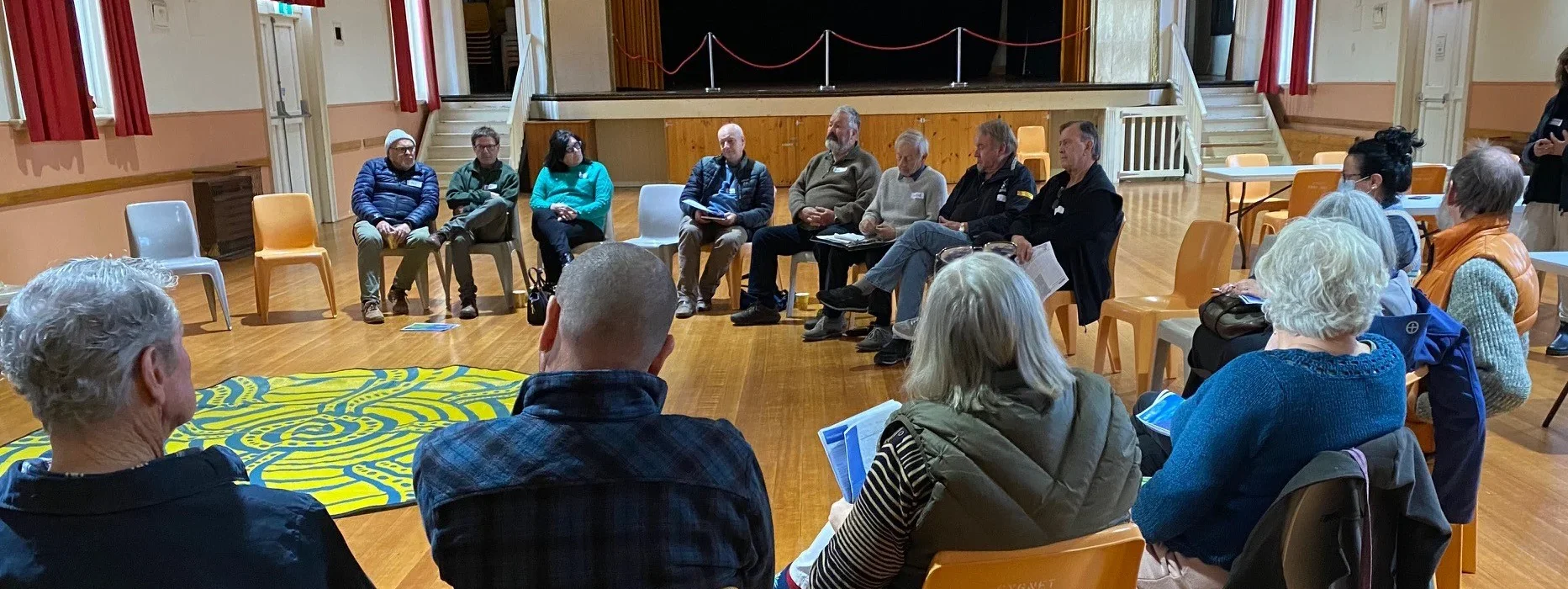 A group of adults sit in a circle of chairs in a well-lit hall with wooden floors, red curtains, and a stage in the background, engaged in a discussion or meeting.