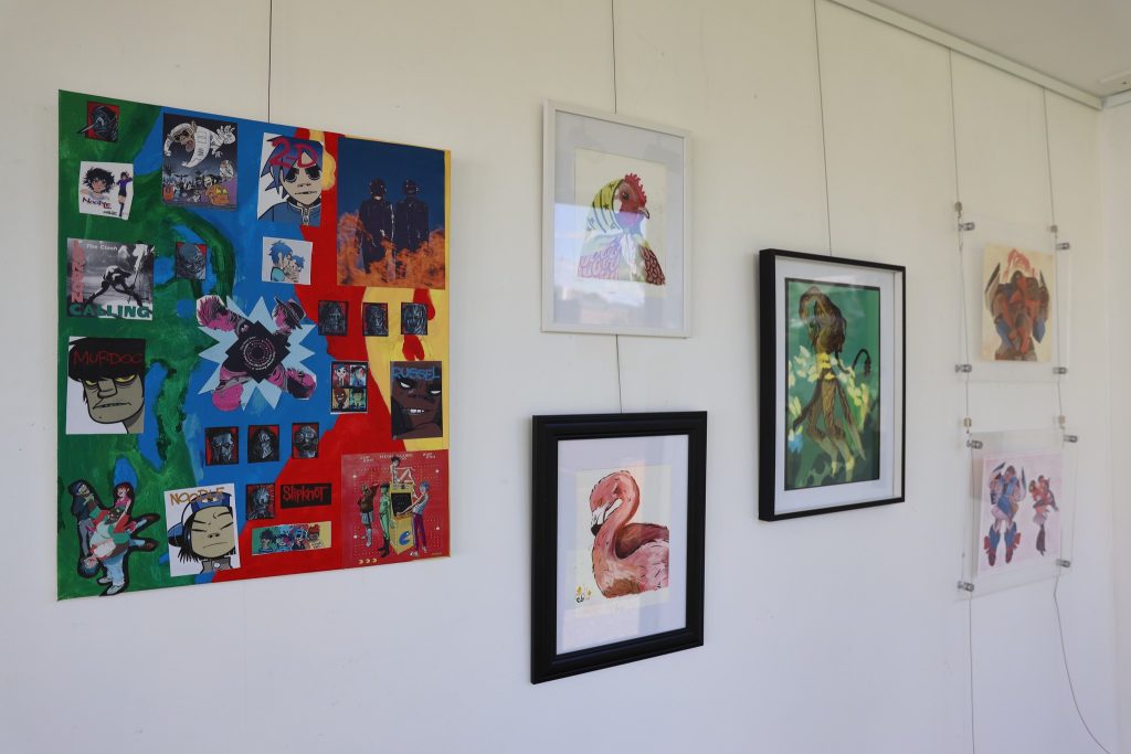 A white gallery wall displays colorful framed artwork, including a collage of album covers, a flamingo illustration, and several abstract and portrait prints arranged in a neat row.
