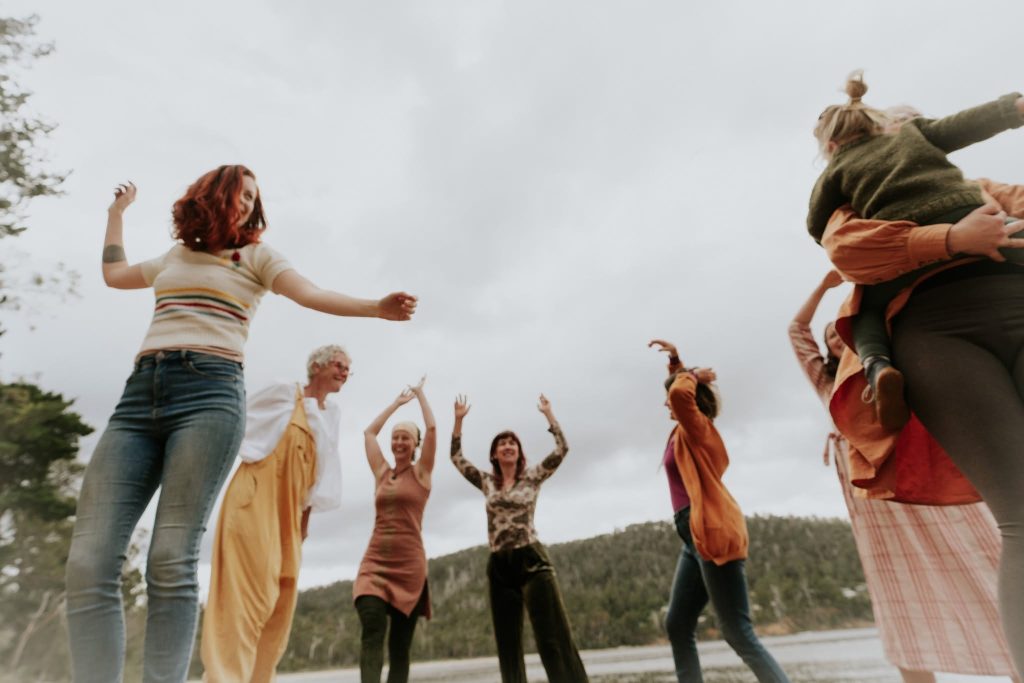 A group of people outdoors cheerfully dancing and raising their arms, with trees and hills in the background under a cloudy sky.