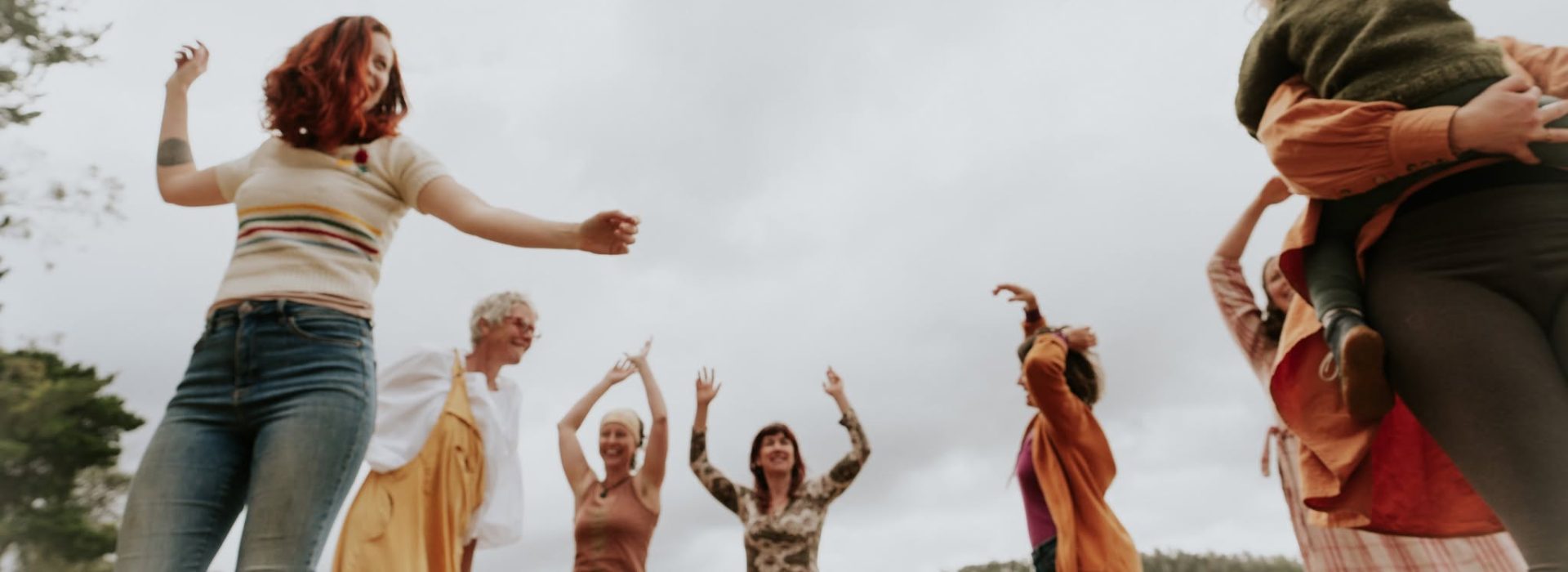 A group of people outdoors cheerfully dancing and raising their arms, with trees and hills in the background under a cloudy sky.