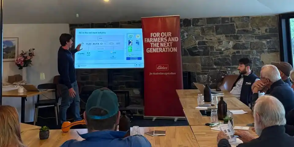 A man stands and points to a presentation projected on a screen in a meeting room, while several people seated at tables listen. A red banner reads For our farmers and the next generation.