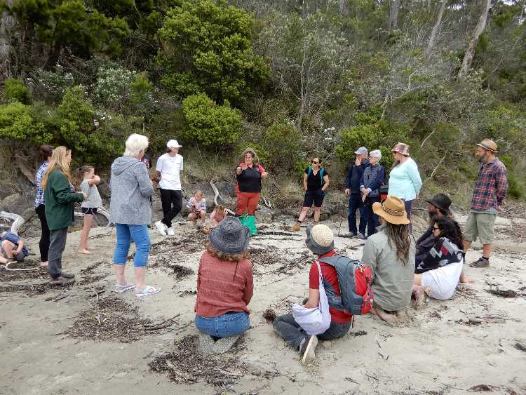 A group of people gather on a sandy beach near forested trees, some standing and some sitting, engaged in conversation or listening to a speaker. The atmosphere appears casual and outdoorsy.