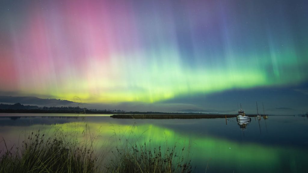 Colorful aurora lights in shades of pink, green, and purple illuminate the night sky over calm water, with a boat anchored near the shore and reflections of the lights on the surface. Grasses frame the foreground.