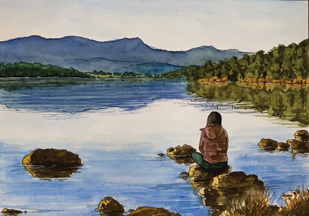 A person sits alone on rocks by the edge of a calm lake, facing away and looking toward distant blue mountains and a forested shoreline under a partly cloudy sky.