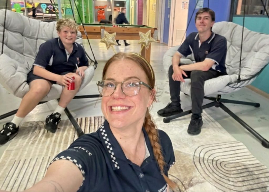 Three people in matching navy shirts sit and smile in a bright, colorful room. One holds a drink, and two sit on cushioned hanging chairs behind the person taking a cheerful selfie. Stars are seen above their heads.