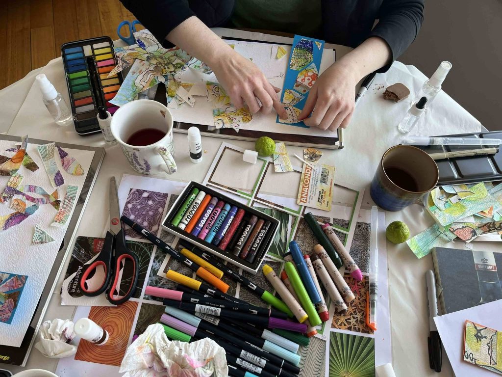 A person creates colorful art at a cluttered table with markers, colored pencils, scissors, paint, scraps of paper, mugs of tea, and various art supplies spread out around their hands.